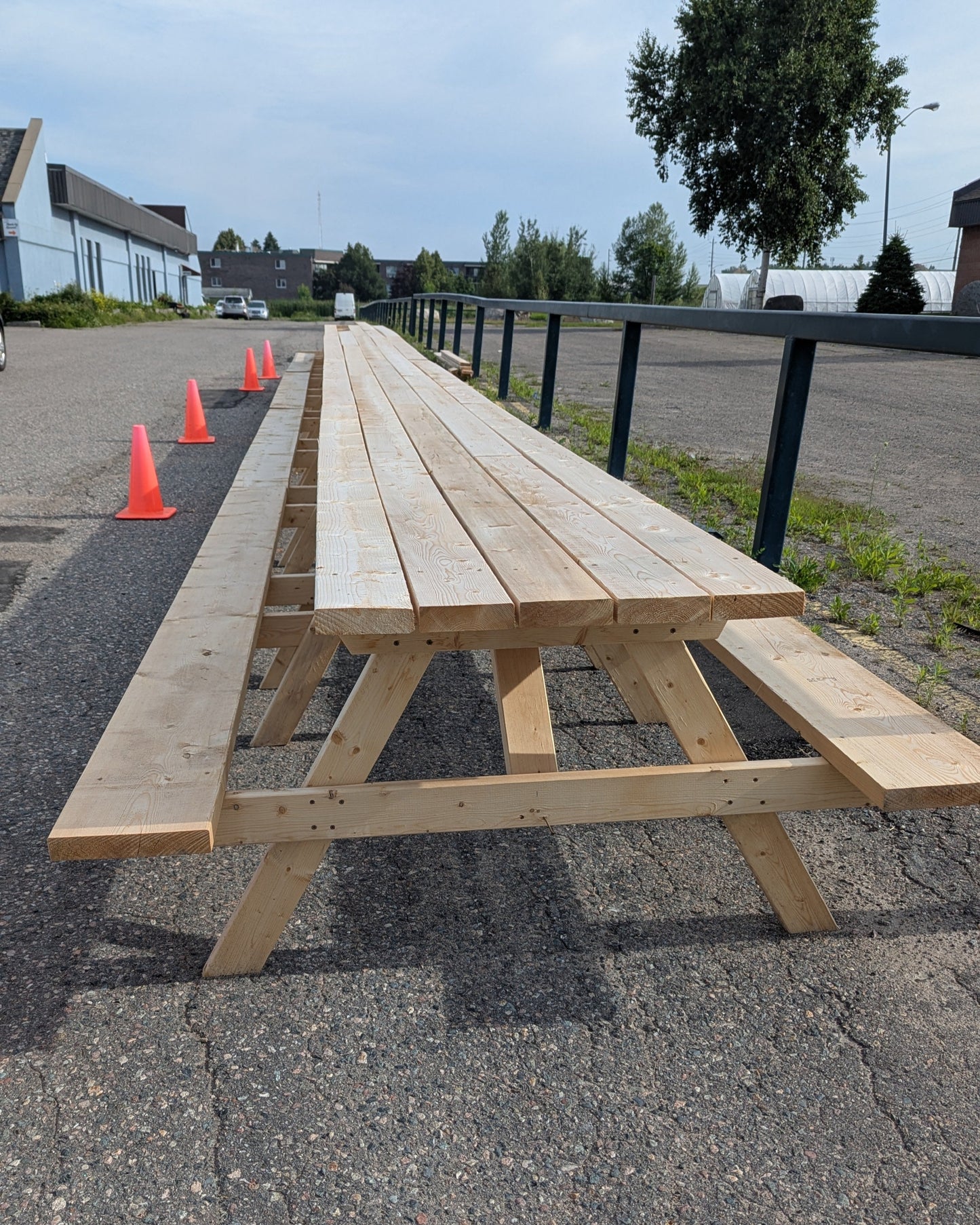 A very long wooden picnic table on a paved surface with traffic cones and a building in the background.