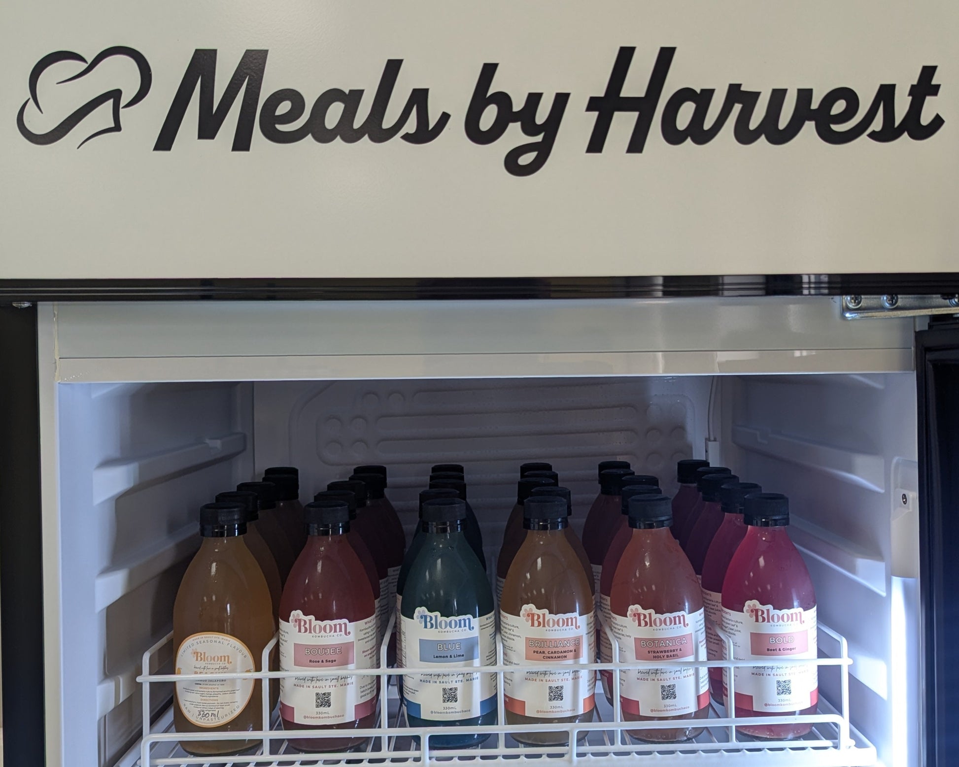 Refrigerator with 'Meals by Harvest' branding and various bottles inside.