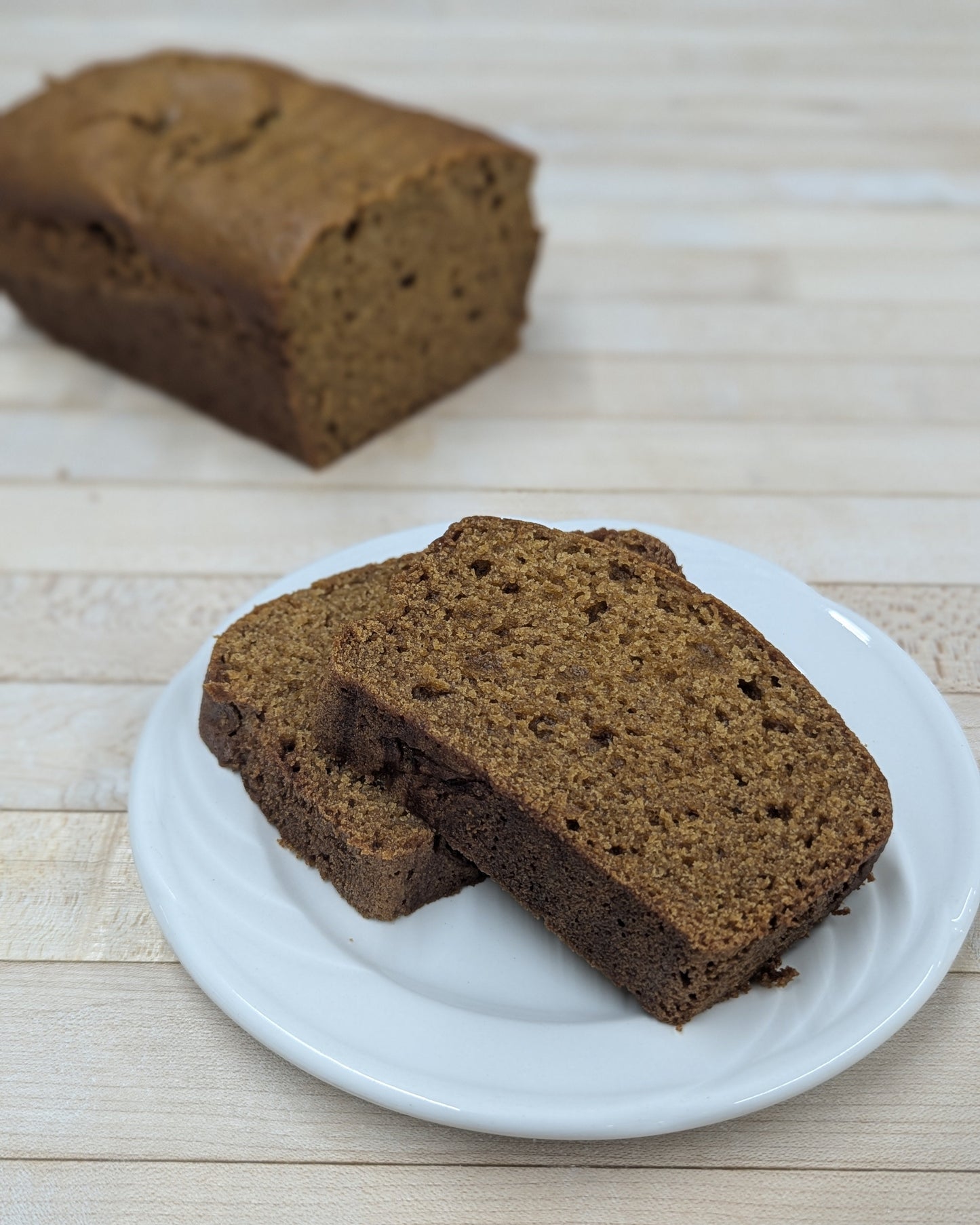 two slices of pumpkin loaf on a white plate sitting on a wooden table, with the rest of the loaf on the table behind it